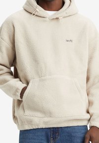 Uomo che indossa un hoodie in pile color crema con tasca frontale e logo "Levi's" sul petto, abbinato a jeans blu.