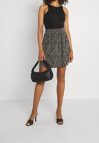 Haut sans manches noir associé à une jupe à motifs, arrivant au genou. Tient un sac à main noir tressé, porte des talons ouverts noirs.