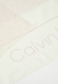 Écharpe de couleur claire avec une texture lisse. Présente un motif subtil de logo entrelacé et des bords à franges. Comprend le nom de la marque imprimé.