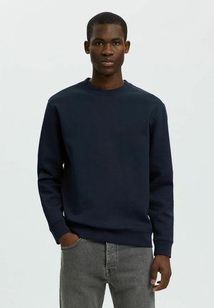 Jeune homme portant un sweat-shirt bleu foncé uni et un jean gris, debout avec une main dans la poche devant un fond blanc uni.
