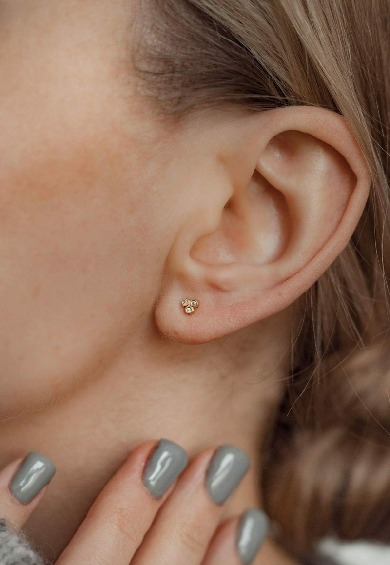 Goldene Steckerohrringe mit drei kleinen Kugeln, zwei polierte Goldkugeln und eine mit einem klaren Stein, getragen am Ohr eines Models neben kurzen grauen Nägeln.