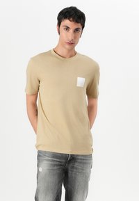 T-shirt en coton beige avec col rond, manches courtes et une étiquette blanche carrée sur la poitrine gauche. Associé à un jean gris déchiré.
