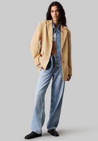 Beżowa marynarka dopasowana na tle niebieskiej koszuli dżinsowej, w zestawieniu z jasnoniebieskimi spodniami o szerokich nogawkach i czarnymi botkami za kostkę. Gładkie faktury i kontrastujące kolory.