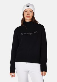 Maglione lavorato a maglia nera con colletto alto e polsini a costine. Presenta la scritta "Rossignal" in argento sul petto. Indossato con pantaloni bianchi.
