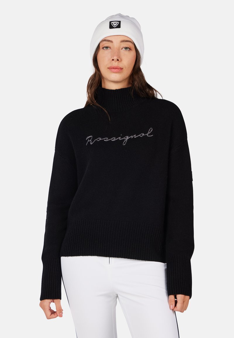 Maglione lavorato a maglia nera con colletto alto e polsini a costine. Presenta la scritta "Rossignal" in argento sul petto. Indossato con pantaloni bianchi.