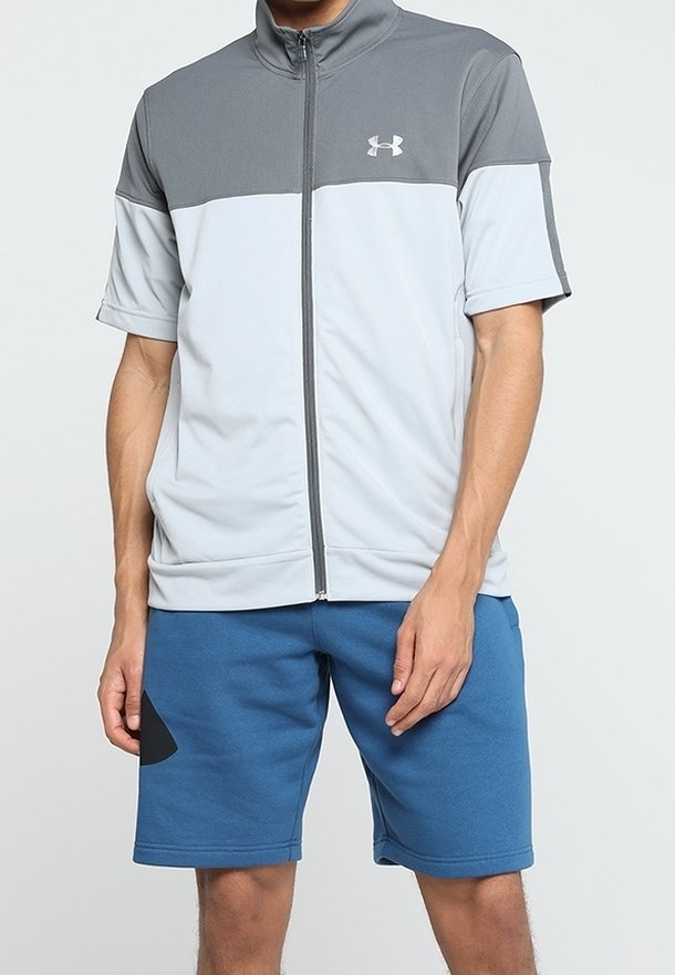 Homme portant une veste zippée à manches courtes grise et blanche et un short bleu avec un motif noir sur la jambe gauche.
