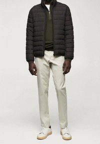 Giacca nera imbottita con colletto alto, maglione verde oliva sotto, pantaloni beige chiaro e sneakers bianche con suola beige.