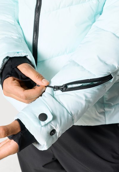 Ανοιχτό μπλε φουσκωτό μπουφάν με τσέπη με φερμουάρ στο μανίκι, διαθέτοντας μαύρα εξαρτήματα και αντίθετα μαύρα εσωτερικά μανίκια.