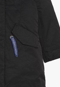 Svart vadderad kappa med en slank design, som har en sned ficka och en blå snörning med silverdetaljer. Slät textur.