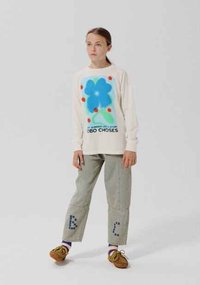 Young person standing, wearing a white sweatshirt with a large blue flower graphic and gray pants with letters on the legs.