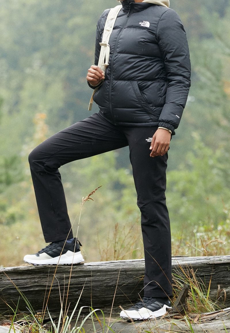 Personne portant une veste et un pantalon noirs The North Face, debout en plein air sur une bûche avec un pied relevé, portant un sac à dos beige.