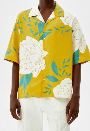 Homme portant une chemise jaune à manches courtes avec de grandes fleurs blanches et des feuilles bleues, associée à un pantalon blanc.