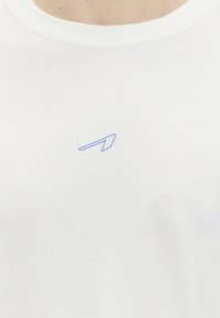 Близък план на бяла тениска с малко, минималистично синьо геометрично лого, центрирано на гърдите.