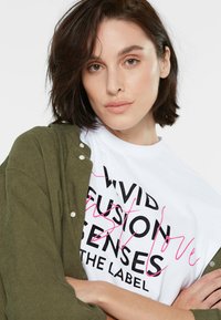 Weißes Grafik-T-Shirt mit schwarzem und pinkem Text, drunter getragen unter einer olivgrünen Strickjacke mit gerippter Textur und weißen Knöpfen.