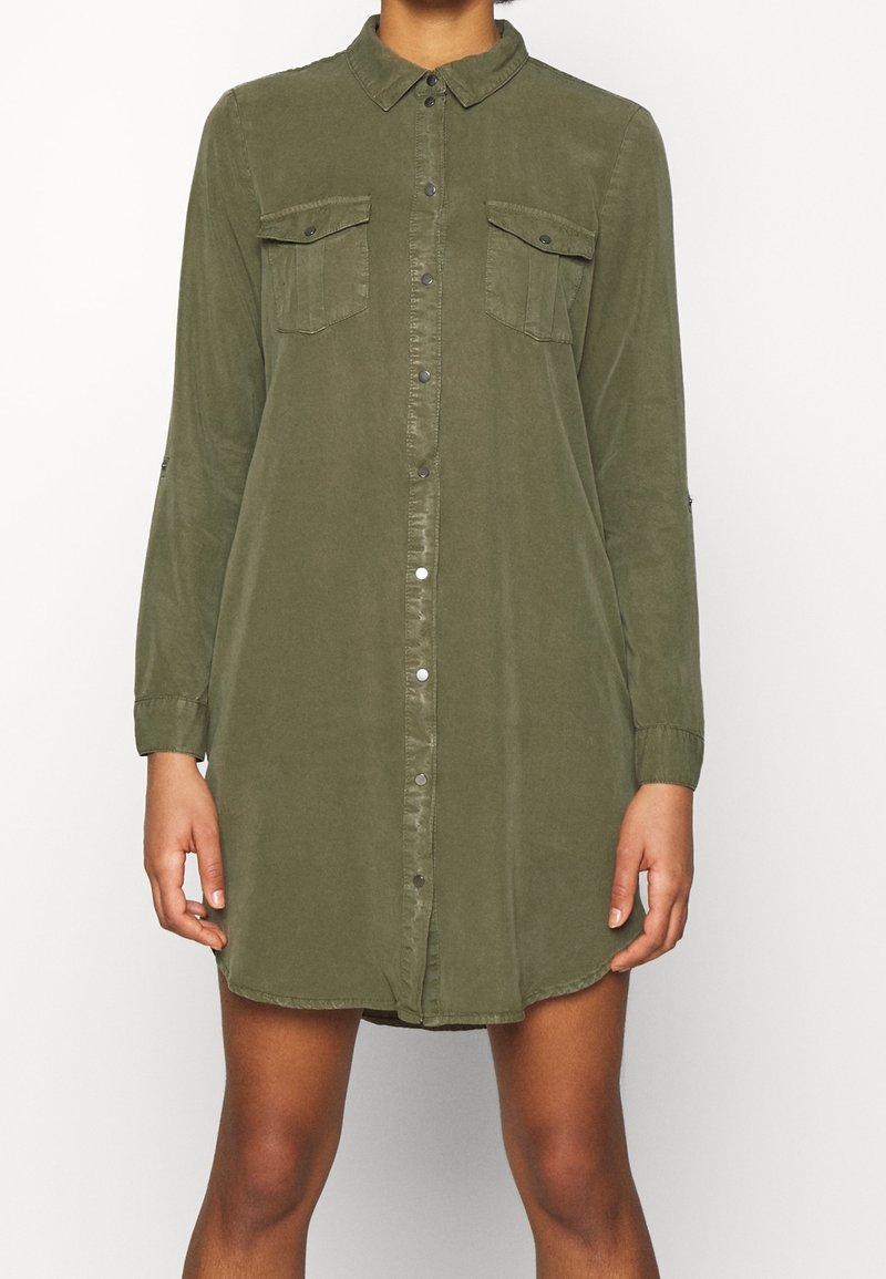 Femme portant une robe-chemise à manches longues couleur olive avec patte de boutonnage devant et poches poitrine à rabat boutonnées.