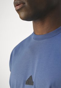 Niebieska koszulka z okrągłym dekoltem, wykonana z miękkiego materiału, z delikatną teksturą. Projekt zawiera małe czarne logo Adidas na przodzie.