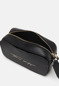 Borsa rettangolare in pelle nera con zip e hardware dorati. Presenta un logo impresso sul davanti e un interno aperto.