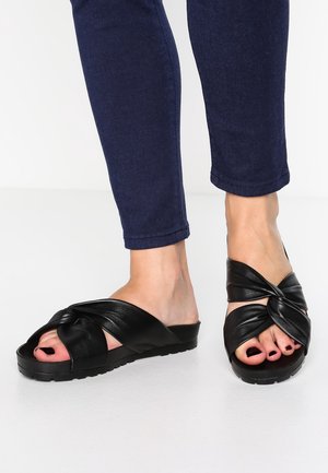 Fødder iført sorte sandaler med snoede stropper, kombineret med mørkeblå slim-fit jeans mod en hvid baggrund.