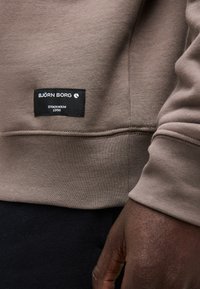 Κοντινό πλάνο του τελειώματος ενός καφέ φούτερ με μια μαύρη ετικέτα Björn Borg και ένα χέρι που ακουμπά κοντά στον γοφό του φορέα.