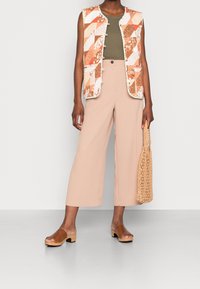 Chaleco de patchwork en tonos naranjas y blancos, top de tirantes oliva, pantalones de pierna ancha beiges, mocasines de tacón de madera marrones y un bolso de mimbre.