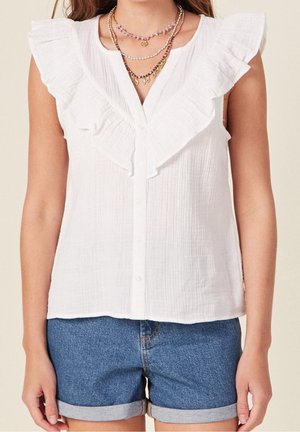Kvinde iført hvid ærmeløs bluse med flæser og knapper samt lagdelte halskæder, kombineret med optrukne denimshorts.