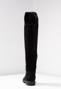 Vysoká černá semišová bota s plochou gumovou podrážkou, stojící vzpřímeně na bílé ploše na bílém pozadí.