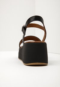 Černé platformové klínové sandály s páskem kolem kotníku a stříbrnou sponou, z pohledu zezadu na bílé podložce a s bílým pozadím.