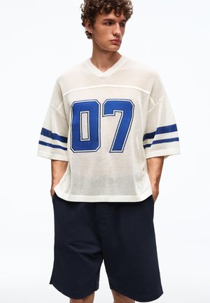 Młody mężczyzna w białej siateczkowej sportowej koszulce z niebieską cyfrą "07" i paskami, w luźnych ciemnych szortach, z rękami w kieszeniach, patrzący na bok.