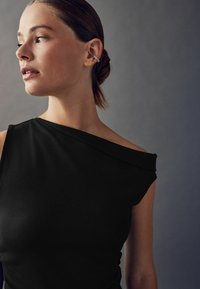 Kicsit hátra fésült hajú nő egy fekete, egyvállú felsőben és ezüst- és aranyfülbevalókkal a fülén, balra nézve egy szürke háttér előtt.