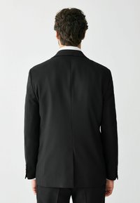 Man met kort krullend haar, gekleed in een zwart pak en wit overhemd, staande met zijn rug naar voren tegen een effen lichte achtergrond.