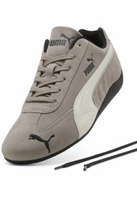 Baskets Puma en suède gris avec bande latérale blanche, semelle noire, logos Puma noirs, et deux lacets noirs détachés à côté de la chaussure sur fond blanc.