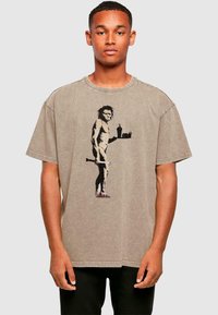 Taubfarbendes, übergroßes Baumwoll-T-Shirt mit einem Motiv eines Höhlenmenschen, der ein Getränk hält. Kurze Ärmel, runder Halsausschnitt und ausgewaschene Textur.