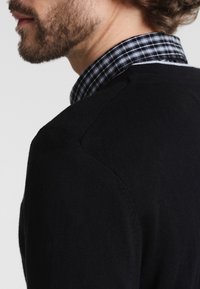 Czarny dziany sweter o gładkiej fakturze, z kołnierzem odwiniętym w dół, pod którym widoczny jest wzór w kratę. Widok od tyłu, pokazujący szew na ramieniu.