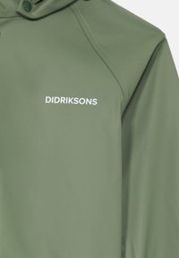 Zelena nepremočljiva jakna z gladko teksturo in zadrgo spredaj. Predstavlja ime blagovne znamke "Didriksons" v beli barvi na predelu prsnega koša.