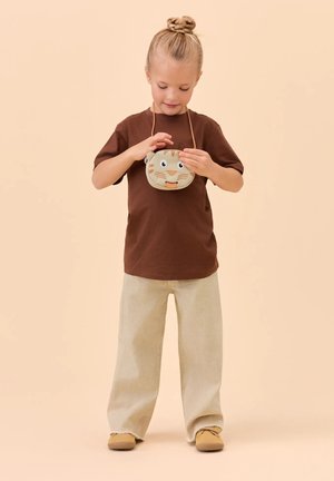 Junges Kind mit blondem Haar in einem Dutt, trägt ein braunes Hemd und beige Hose, hält vor sich eine kleine Tasche mit Tigerkopf-Motiv.