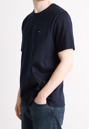 Mężczyzna ubrany w gładki granatowy T-shirt z krótkim rękawem i małym logo na piersi, niebieskie dżinsy, jedną rękę w kieszeni, na białym tle.