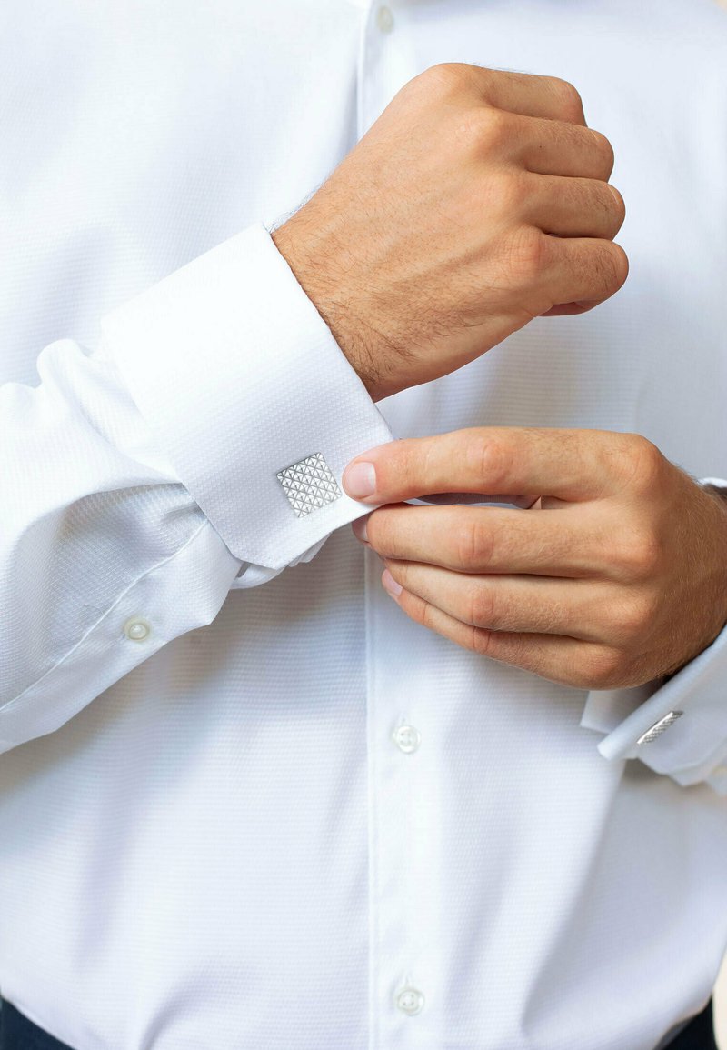 Chemise blanche en tissu texturé. Bouton de manchette carré en argent sur une manche en cours d'ajustement. Boutons visibles le long du devant de la chemise.