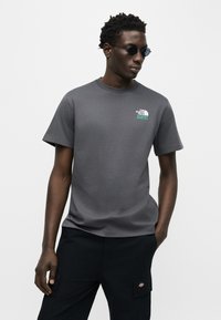 Άνδρας που φοράει στρογγυλά μαύρα γυαλιά ηλίου, σκούρο γκρι μπλουζάκι The North Face και μαύρο παντελόνι με το ένα χέρι στην τσέπη, στέκεται μπροστά σε λευκό φόντο.