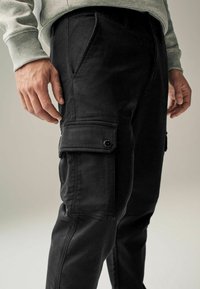 Mustat cargo-housut denimmateriaalista, joissa on sivutaskut napin sulkemisella, kapea malli ja hieman teksturoitu pinta.