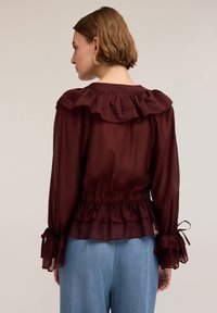 Blusa bordeaux con scollo arricciato e vita gather. Tessuto trasparente con maniche lunghe che presentano decorazioni legate ai polsini.