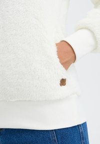 Balta, purus megztinis su minkšta tekstūra, turintis banguotus rankogalius ir apačią. Ant šono yra mažas rudas žymeklis su "xm" detale.