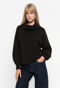 Blonde Frau, die einen schwarzen, grob gestrickten Rollkragenpullover und dunkelblaue Jeans trägt, mit einer Hand in ihrer Tasche, stehend vor einem weißen Hintergrund.