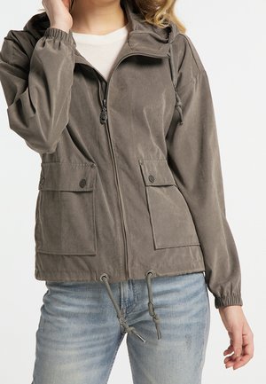 Femme portant une veste grise zippée avec poches à rabat à l'avant et cordons, associée à un jean bleu clair sur fond blanc.