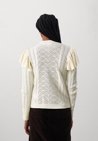 Maglione crema con motivi a diamante testurizzati, maniche arricciate e fondo a costine. La vista posteriore mette in risalto il design lavorato con dettagli.