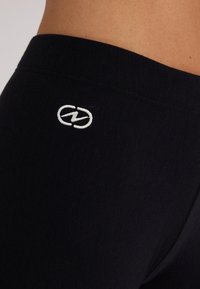 Zwarte leggings met een elastische tailleband, met een klein wit logo geborduurd op de achterkant. De stof lijkt glad en gestructureerd.