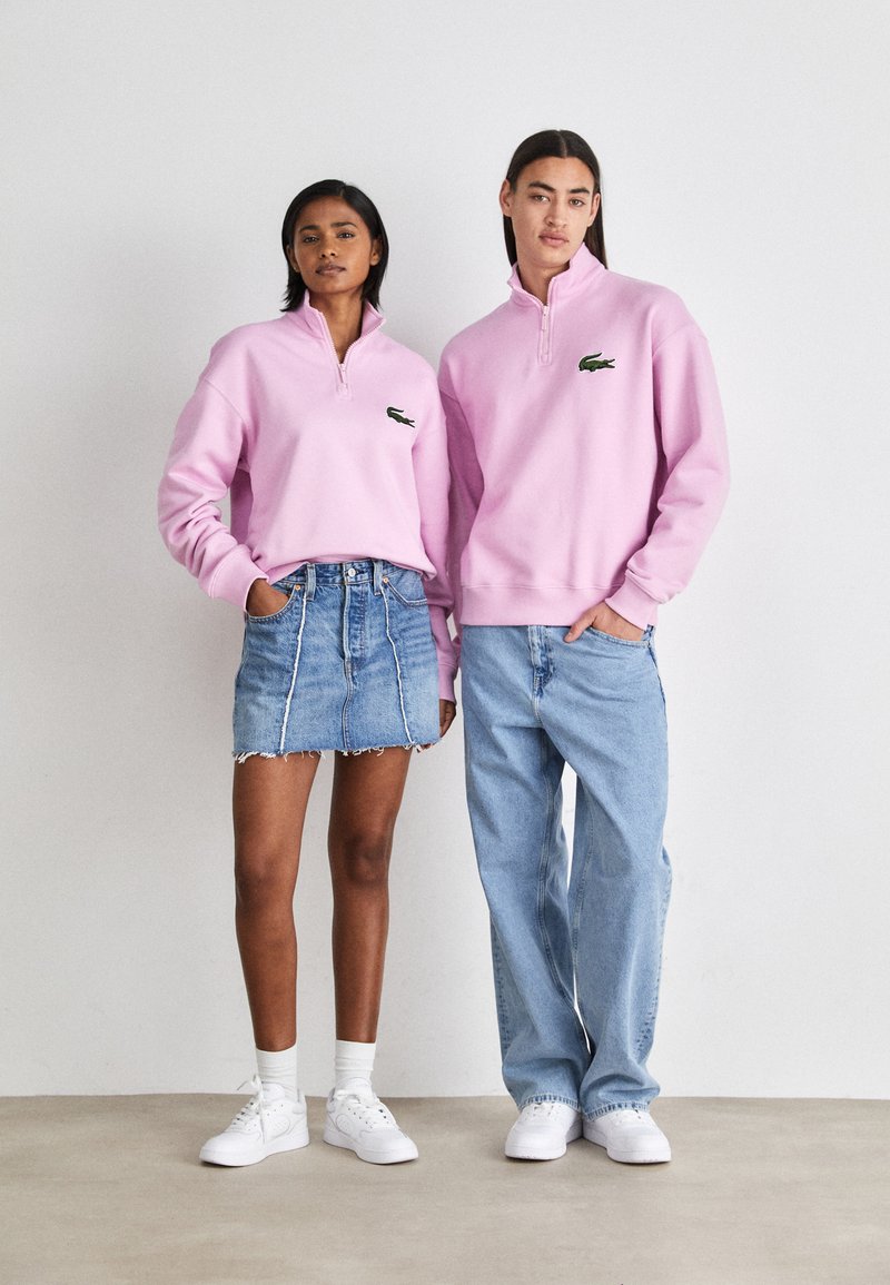 Felpe rosa con zip e logo verde; abbinate a una gonna di jeans e jeans loose azzurri; entrambi indossano sneakers bianche.