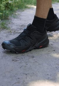 Schwarze Laufschuhe aus strukturiertem Stoff, mit geformten Akzenten und roten Details. Der Schuh verfügt über eine robuste Sohle und ein sicheres Schnürsystem.