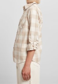 Donna che indossa una camicia a maniche lunghe beige e bianca a scacchi con le maniche arrotolate e pantaloni chiari, mostrata di profilo su uno sfondo semplice.