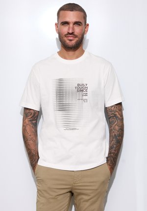 Homme aux cheveux courts et tatouages sur toute la manche, portant un t-shirt blanc à motif graphique et un pantalon beige, debout les mains dans les poches.