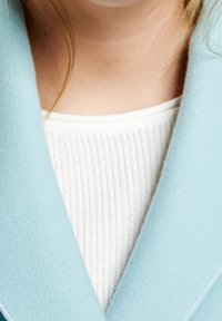 Un cuello de lana de color azul claro con una textura suave, superpuesto a un suéter blanco de canalé. La costura visible acentúa los bordes del cuello.